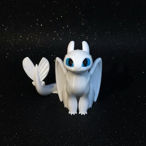 Puede incluir: Una figura de dragón blanco con orejas grandes y puntiagudas y ojos de color azul brillante. La criatura tiene un acabado liso y brillante, alas plegadas y una cola larga con una aleta en forma de corazón en el extremo y garras.