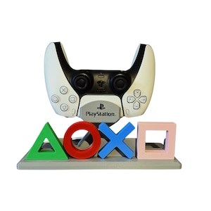 Op de afbeelding: Een witte en zwarte PlayStation 5-controller staat boven een grijze basis met kleurrijke gamecontrollersymbolen. De basis heeft een groene driehoek, een rode cirkel, een blauwe X en een roze vierkant. Het PlayStation-logo is zichtbaar.