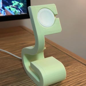 Puede incluir: Un soporte de carga verde claro para Apple Watch. El soporte tiene un diseño curvo con una plataforma de carga circular en la parte superior. Un cable de carga blanco está conectado al soporte. El soporte está hecho de un material liso y mate.