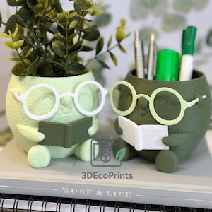 Puede incluir: Dos macetas originales, una verde claro y otra verde oscuro, con forma de personajes que llevan gafas y sostienen un libro. Una contiene una planta, la otra bolígrafos y rotuladores. Las macetas están sobre un cuaderno con el texto "WORK & LIFE". El logotipo "3DEcoPrints" es visible.