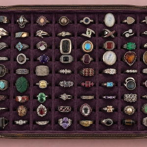 May include: A rectangular jewelry display tray with a dark purple velvet lining and a decorative bronze-toned frame, containing an organized collection of eighty assorted rings featuring various gemstones, metal bands, and intricate settings.