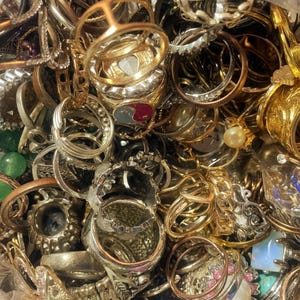 May include: A large collection of assorted rings in various metal tones including gold, silver, and copper. The pile features rings with diverse designs, some containing embedded pearls, colorful stones, and intricate metalwork patterns.