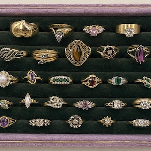 May include: A collection of twenty-five assorted rings displayed in a rectangular tray with a purple frame and dark green velvet slots. The rings feature various metal tones, including gold and silver, set with diverse gemstones like pearls, diamonds, and colored stones.