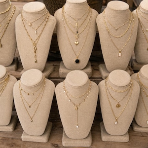 May include: Ten beige fabric necklace display busts, each showcasing a layered gold-tone chain necklace set. The necklaces feature various pendants including hearts, coins, geometric shapes, and charms, with different chain styles like rope and link.