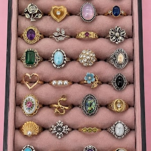May include: A collection of twenty-eight assorted rings displayed in a rectangular jewelry organizer with a pink velvet interior. The rings feature various designs including gemstones, floral motifs, hearts, celestial shapes, and metal bands in gold and silver tones.