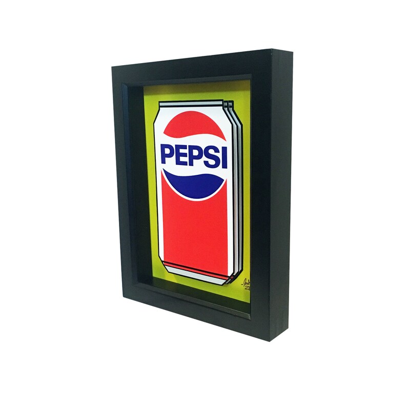 May include: A black framed print of a yellow background with a red and blue Pepsi can.