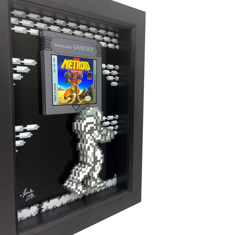 May include: A framed 3D pixel art piece featuring a gray and white pixelated character from the video game Metroid. The character is holding a gun and is standing on a platform. The frame is black and has a glass front. The game cartridge for Metroid is displayed in the frame.