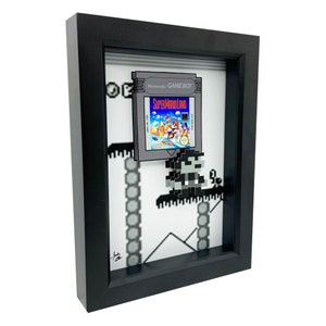 May include: A framed 3D pixel art piece featuring a Super Mario Land Game Boy cartridge and a pixelated Mario character. The artwork is set against a white background with a pixelated background of a level from the game.
