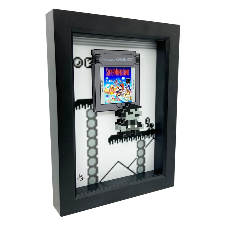 May include: A framed 3D pixel art piece featuring a Super Mario Land Game Boy cartridge and a pixelated Mario character. The artwork is set against a white background with a pixelated background of a level from the game.
