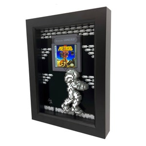May include: A framed 3D pixel art piece featuring the classic Nintendo Game Boy game Metroid. The frame is black and the artwork is in a retro style, with a pixelated image of Samus Aran, the protagonist of the game, walking across a platform. The game cartridge is displayed in the frame, with the title "Metroid" visible.