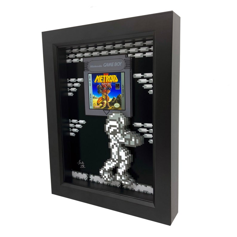 May include: A framed 3D pixel art piece featuring the classic Nintendo Game Boy game Metroid. The frame is black and the artwork is in a retro style, with a pixelated image of Samus Aran, the protagonist of the game, walking across a platform. The game cartridge is displayed in the frame, with the title "Metroid" visible.