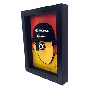 May include: A framed illustration of a character from the movie "A Clockwork Orange". The character is wearing a black hat with the words "A Clockwork Orange" printed on it. The character's face is yellow and has one large eye. The background is red, yellow, and black.