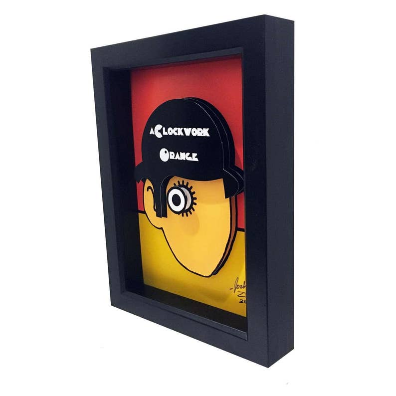 May include: A framed illustration of a character from the movie "A Clockwork Orange". The character is wearing a black hat with the words "A Clockwork Orange" printed on it. The character's face is yellow and has one large eye. The background is red, yellow, and black.