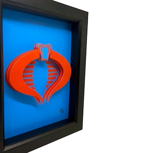 Puede incluir: Una pieza de arte en papel 3D enmarcada del s&iacute;mbolo de Cobra de la caricatura de G.I. Joe. El s&iacute;mbolo es rojo y est&aacute; en capas para crear un efecto 3D. El fondo es azul.
