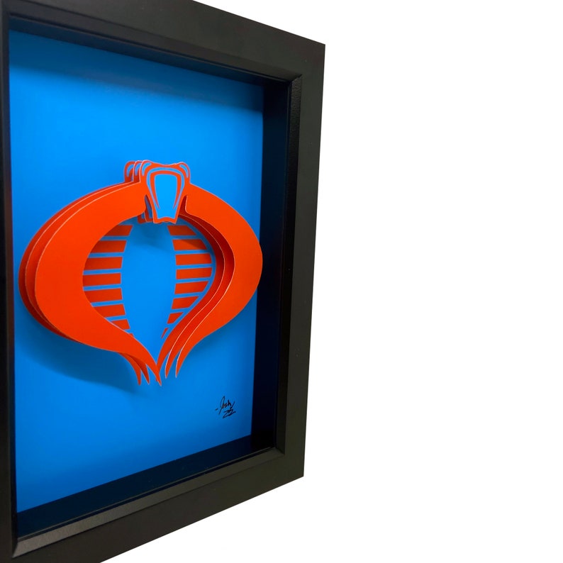 Puede incluir: Una pieza de arte en papel 3D enmarcada del s&iacute;mbolo de Cobra de la caricatura de G.I. Joe. El s&iacute;mbolo es rojo y est&aacute; en capas para crear un efecto 3D. El fondo es azul.