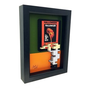 May include: A framed 3D pixel art piece featuring the iconic "Halloween" movie poster. The poster is in a black frame with a green and orange mat. The pixel art is of Michael Myers, the film's antagonist, holding a knife. The piece is signed "Jack 2022".