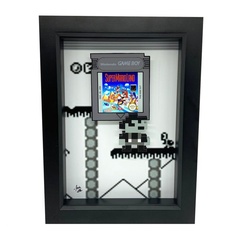 May include: A framed 3D pixel art piece featuring a Super Mario Land Game Boy cartridge and a pixelated Mario character. The background is a pixelated level from the game.