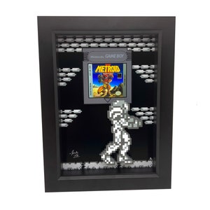 May include: A framed 3D pixel art piece featuring the classic Nintendo Game Boy game Metroid. The game cartridge is displayed in a black frame with a pixelated background. A pixelated character from the game is walking across the bottom of the frame.