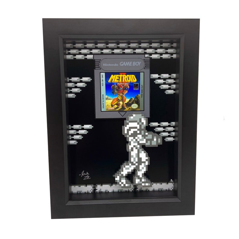 May include: A framed 3D pixel art piece featuring the classic Nintendo Game Boy game Metroid. The game cartridge is displayed in a black frame with a pixelated background. A pixelated character from the game is walking across the bottom of the frame.