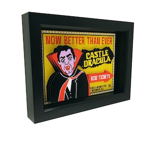 May include: A framed illustration of a vintage amusement park ride advertisement. The illustration features a vampire with a red cape and the text "Now Better Than Ever Castle Dracula Ride Tickets."