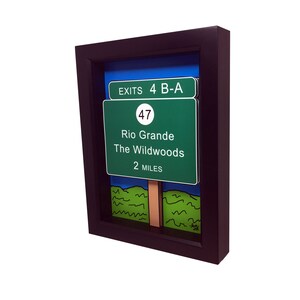 May include: A framed artwork featuring a green highway sign with the text "EXITS 4 B-A", "47", "Rio Grande", "The Wildwoods", and "2 MILES". The sign is set against a blue sky and green grass background.