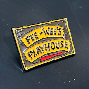 May include: A yellow and black enamel pin with the text "Pee-wee's Playhouse" and a red arrow pointing to the right.