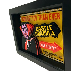 May include: A framed illustration of a vintage amusement park ride called "Castle Dracula". The illustration features a cartoon vampire with a red cape and a yellow background with white polka dots. The text "Now Better Than Ever" is at the top of the illustration. The text "Ride Tickets" is on a red arrow pointing to the right. The text "Nickels' Midway Pier Wildwood, N.J." is at the bottom of the illustration.