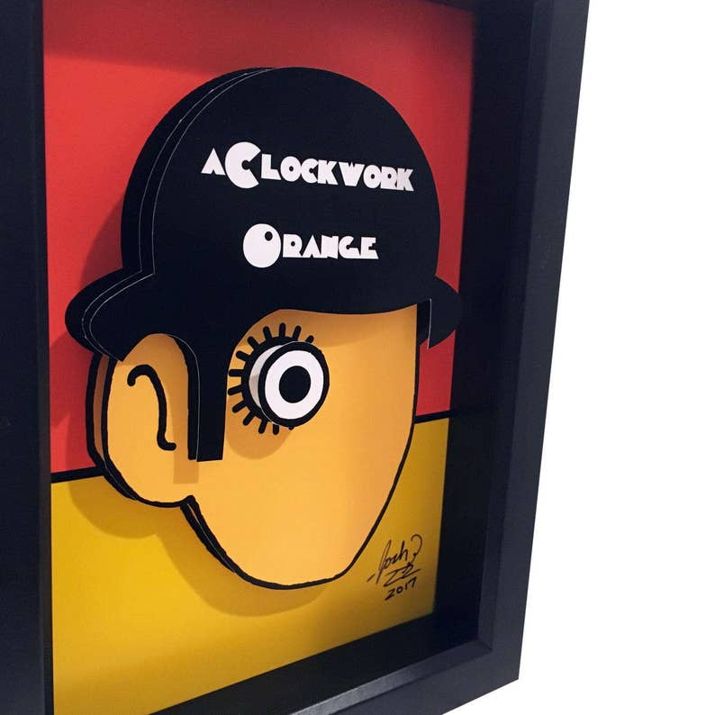 May include: A framed paper art piece featuring a black and white illustration of a character from the movie "A Clockwork Orange". The character has a large eye and a distinctive hairstyle. The artwork is signed and dated "2017".