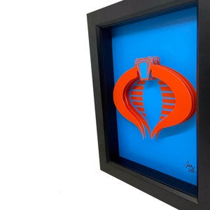 Puede incluir: Una pieza de arte en papel 3D enmarcada del logotipo de Cobra de la caricatura de G.I. Joe. El logotipo es rojo y blanco sobre un fondo azul. El marco es negro.