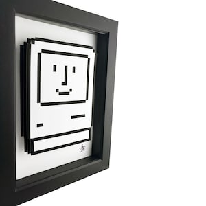 May include: A black and white pixel art print of a vintage Apple computer in a black frame. The computer has a smiling face.