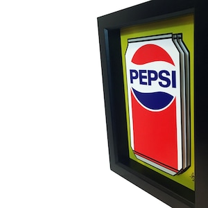 May include: A framed print of a Pepsi can with a yellow background. The can is red and white with the Pepsi logo in blue.