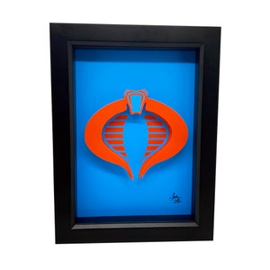 Puede incluir: Una pieza de arte en papel 3D enmarcada del logotipo de Cobra de G.I. Joe. El logotipo es naranja y azul y est&aacute; sobre un fondo azul. El marco es negro.