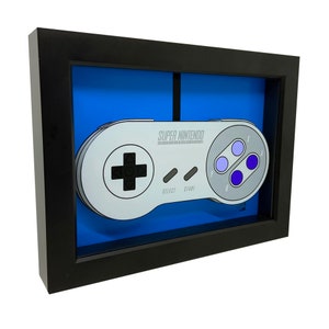May include: Framed artwork featuring a Super Nintendo controller design. The controller is gray with purple and black buttons, set against a blue background. The frame is black, creating a contrast that highlights the controller's details. The text "SUPER NINTENDO" is visible.