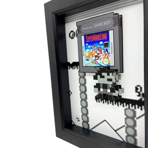 May include: A framed 3D pixel art piece featuring a Super Mario Land Game Boy cartridge and a pixelated Mario character. The cartridge is in a black frame with a white background. The pixel art is on a white background with black lines.
