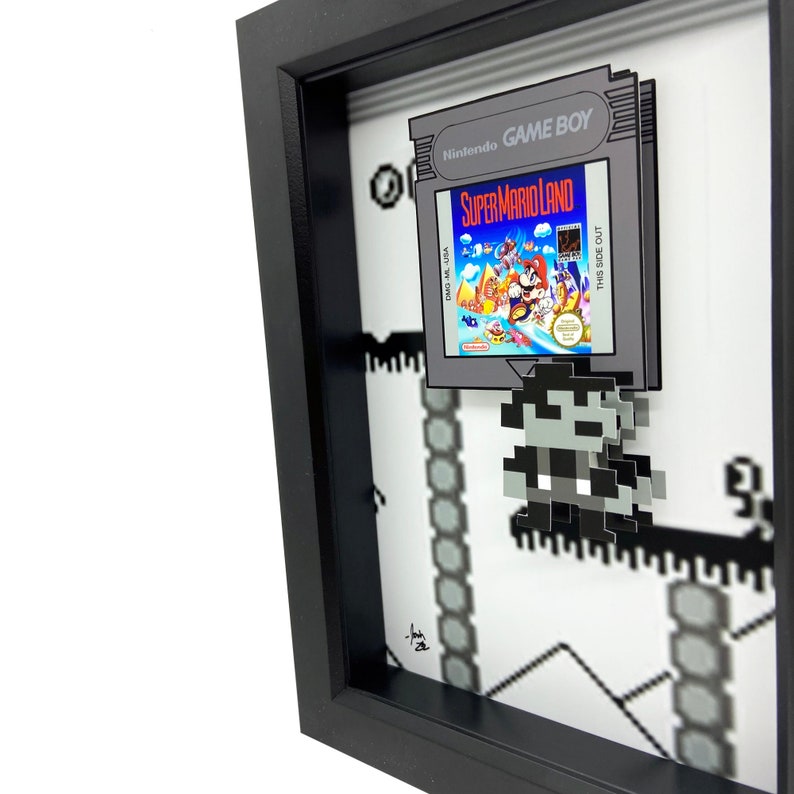 May include: A framed 3D pixel art piece featuring a Super Mario Land Game Boy cartridge and a pixelated Mario character. The cartridge is in a black frame with a white background. The pixel art is on a white background with black lines.