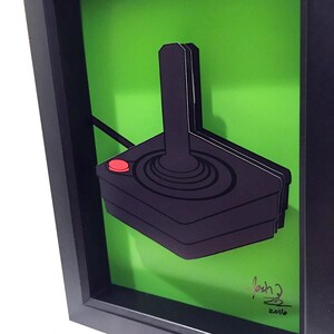 Puede incluir: Un mando de videojuegos vintage de joystick negro y rojo en un marco verde con una firma y la fecha 2016.