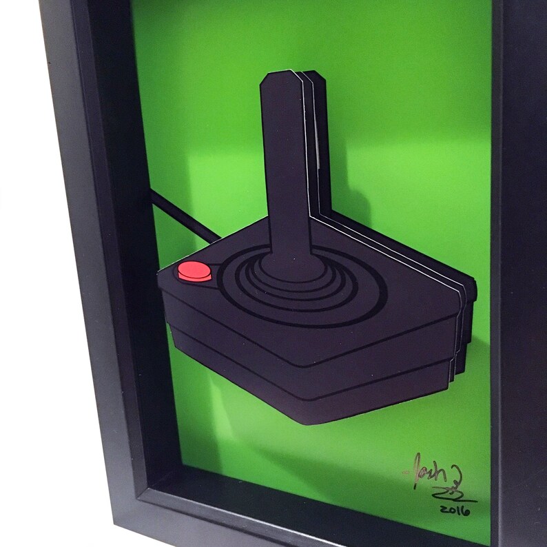 Puede incluir: Un mando de videojuegos vintage de joystick negro y rojo en un marco verde con una firma y la fecha 2016.
