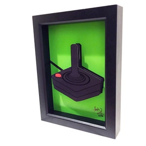 Puede incluir: Un joystick negro y rojo en un marco negro con fondo verde. El joystick es una pieza de arte en papel 3D. La firma del artista y el a&ntilde;o 2016 son visibles en la esquina inferior derecha.