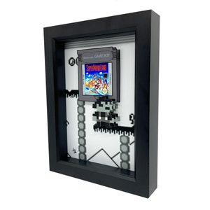 May include: A framed 3D pixel art piece featuring a Super Mario Land Game Boy cartridge and a pixelated Mario character. The frame is black with a white mount and the background is a pixelated level from the game.