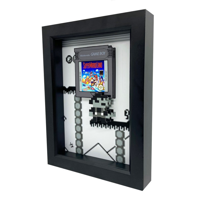 May include: A framed 3D pixel art piece featuring a Super Mario Land Game Boy cartridge and a pixelated Mario character. The frame is black with a white mount and the background is a pixelated level from the game.