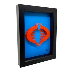 Puede incluir: Una pieza de arte en papel 3D enmarcada del s&iacute;mbolo de Cobra de la caricatura de G.I. Joe. El s&iacute;mbolo es rojo y est&aacute; sobre un fondo azul.
