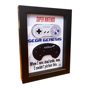 May include: A framed print featuring a black and white illustration of a Super Nintendo and Sega Genesis controller. The print includes the text "SUPER NINTENDO", "SEGA GENESIS", and "When I was dead broke, man, I couldn't picture this."