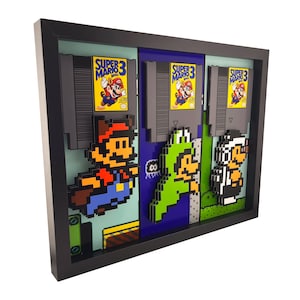 May include: A framed 3D art piece featuring pixelated characters and Super Mario 3 game cartridges. The artwork includes Mario, Luigi, and a character in a black and white outfit. The frame is black, and the background is a mix of blue, green, and gray.