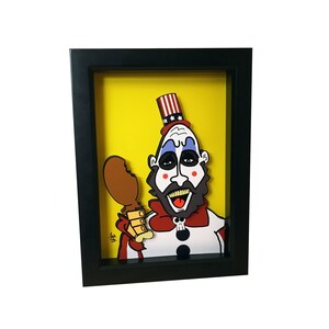May include: A framed illustration of a cartoon character with a red, white, and blue hat, a white beard, and a blue and white face. The character is holding a brown chicken leg and wearing a red bow tie and a white shirt with black dots. The background is yellow.
