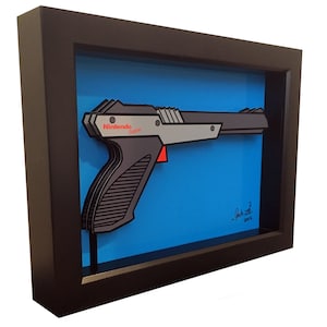 May include: A framed illustration of a Nintendo Zapper light gun, a classic video game accessory. The illustration is in a black frame with a blue background. The Zapper is depicted in a 3D style, with the text "Nintendo Zapper" visible on the gun. The artist's signature and the year 2015 are also visible in the bottom right corner of the frame.