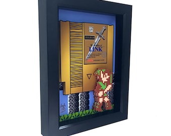 May include: A framed 3D artwork featuring a pixelated Link from the Legend of Zelda video game. The artwork includes a replica of the NES game cartridge for The Legend of Zelda, with a sword sticking out of the top. The artwork is set against a blue and green background.