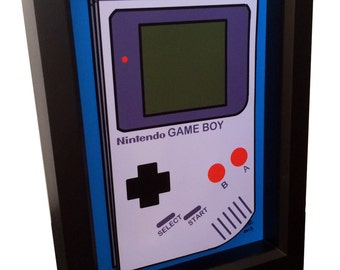 May include: A framed print of a Nintendo Game Boy, a classic handheld video game console. The print is in a black frame with a blue mat. The Game Boy is white with a purple screen and red buttons.