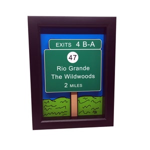 May include: A framed green and white road sign with the text "EXITS 4 B-A" and "Rio Grande The Wildwoods 2 MILES". The sign also has a white circle with the number "47" inside.