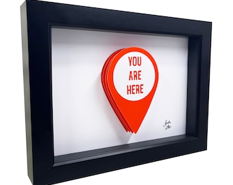Puede incluir: Una imagen enmarcada en negro con un alfiler de mapa de papel 3D rojo que dice "You Are Here" en texto blanco. El alfiler está en capas y tiene un efecto de sombra.