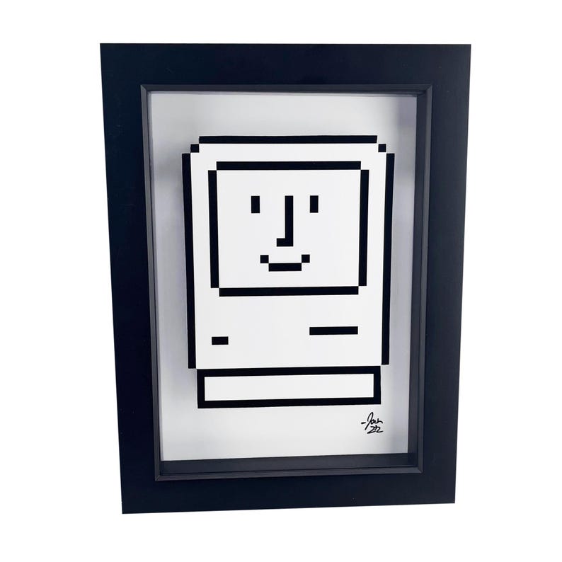 May include: A black framed print of a pixelated image of a vintage Macintosh computer with a smiling face. The print is signed "Josh 92".